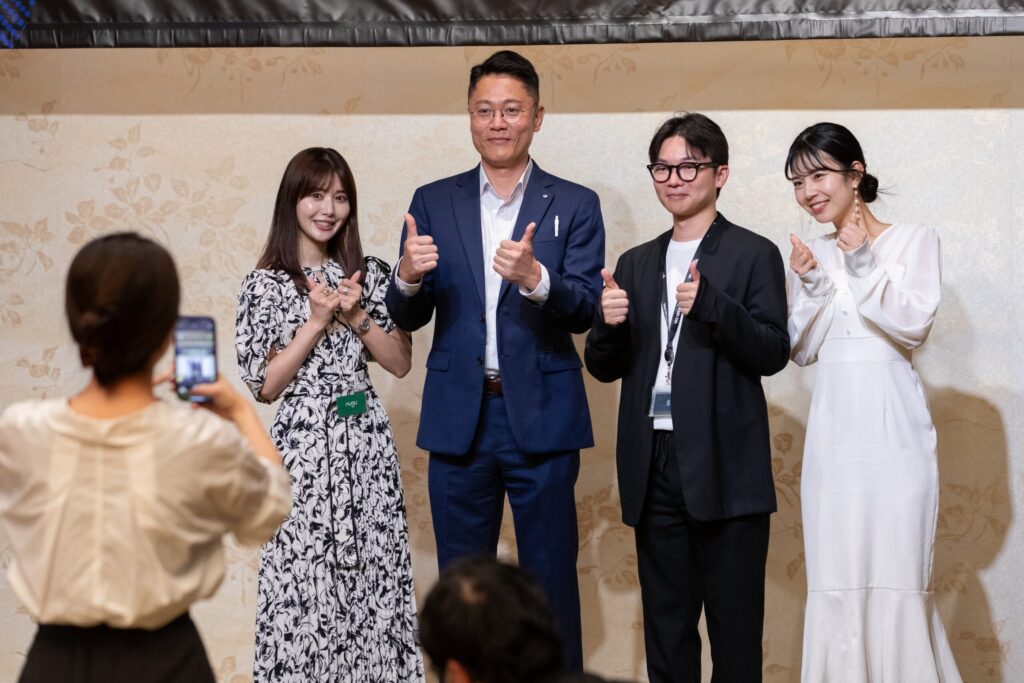 ステージの上で親指を立ててポーズをとるゲストと、スマートフォンで撮影する参加者